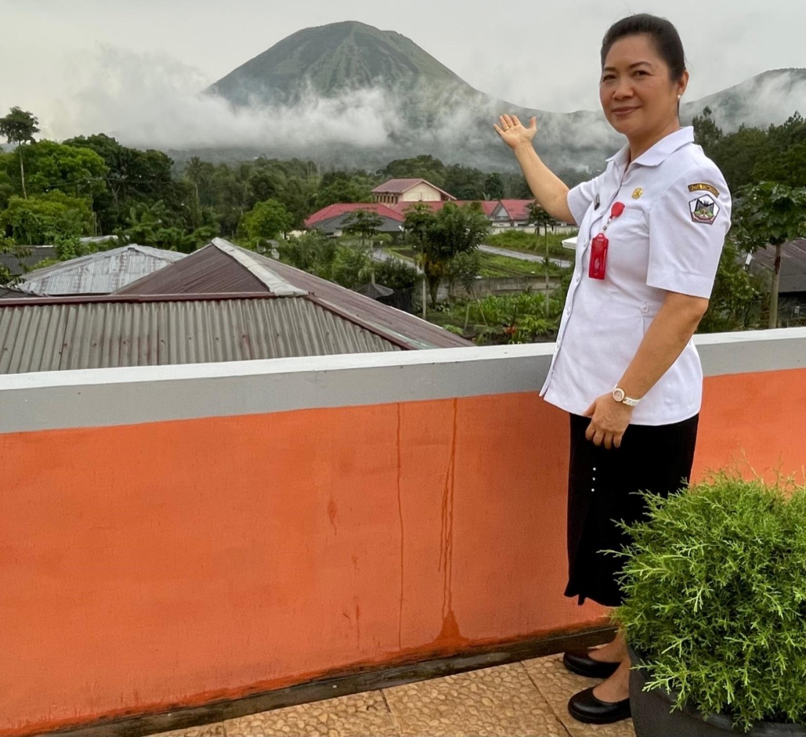 A woman wearing an official government uniform points out a volcanic Mountain Lokon from a rooftop.