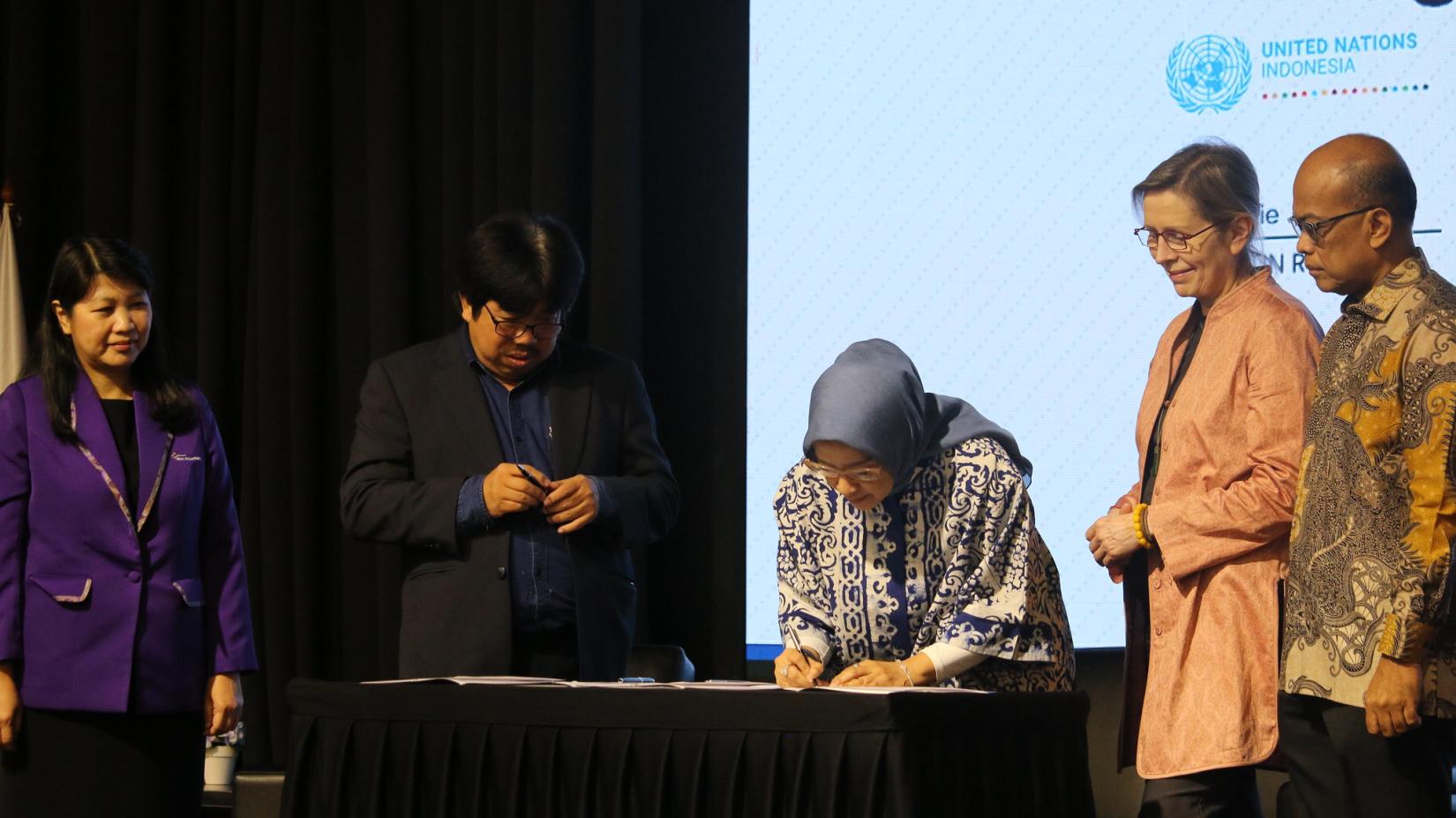 One person signs a paper on a table while four other people witnessing the signing ceremony