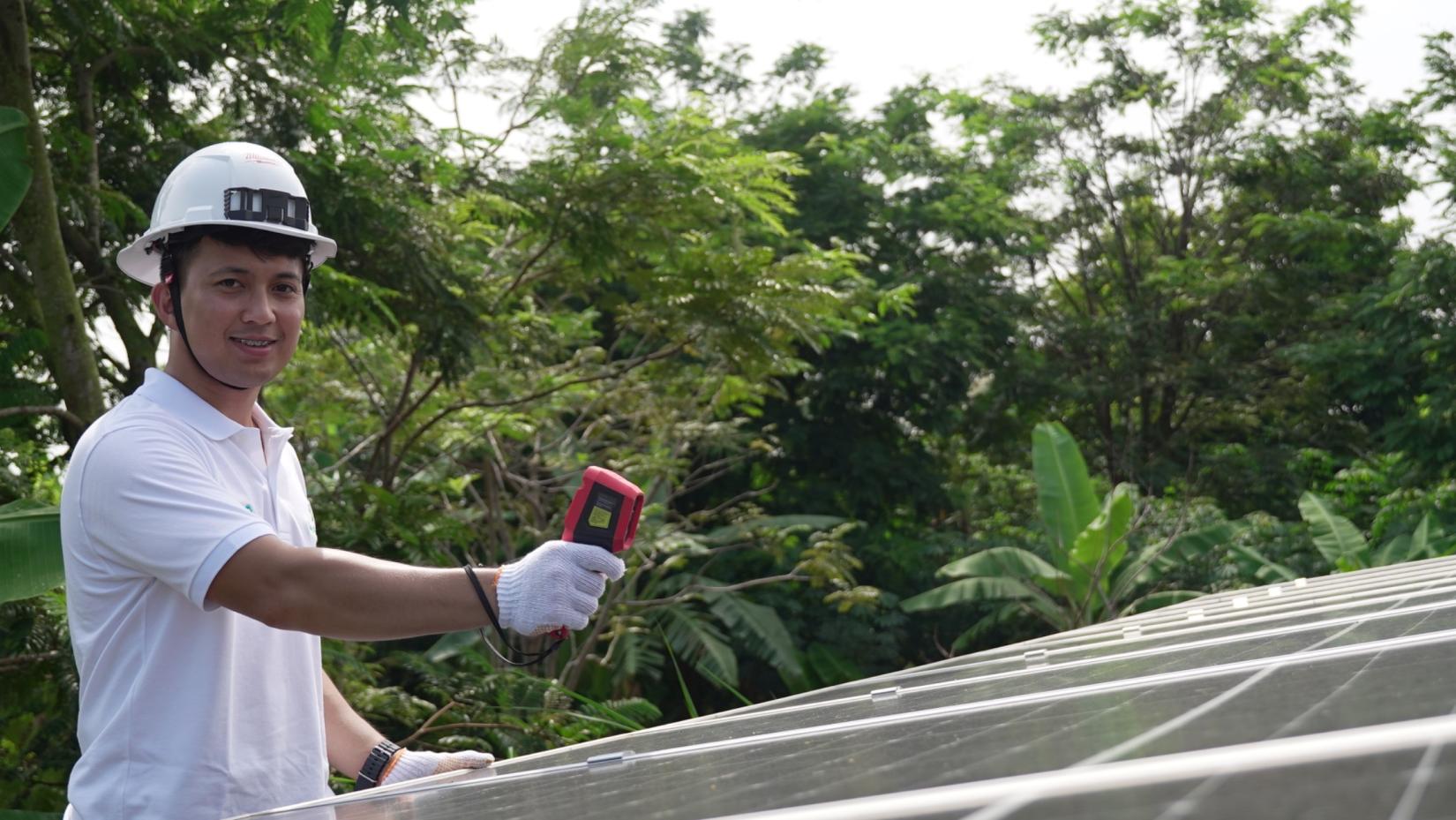 Seorang pria mengenakan helm pengaman putih, kaos polo putih, dan sarung tangan kerja memegang alat pengukur berwarna merah sambil memeriksa panel surya di luar ruangan, dengan latar belakang pepohonan hijau yang rimbun.