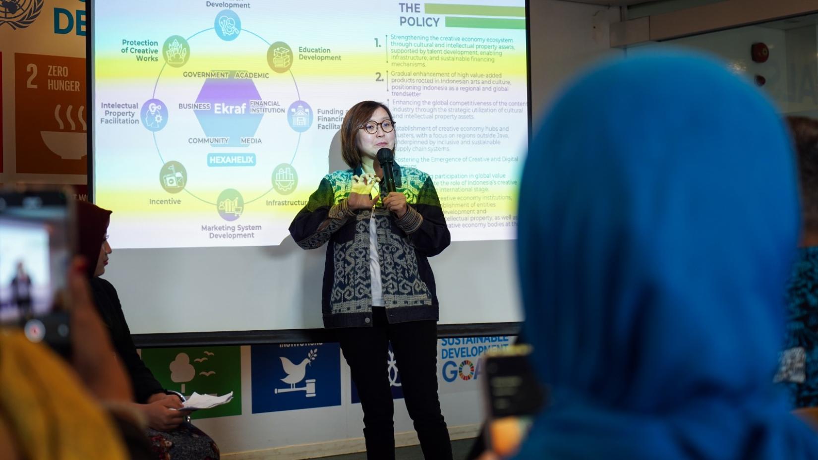 Seorang perempuan berbicara sambil memegang mikrofon di depan layar presentasi yang menampilkan diagram ekosistem ekonomi kreatif, dengan peserta menyimak dari kursi.