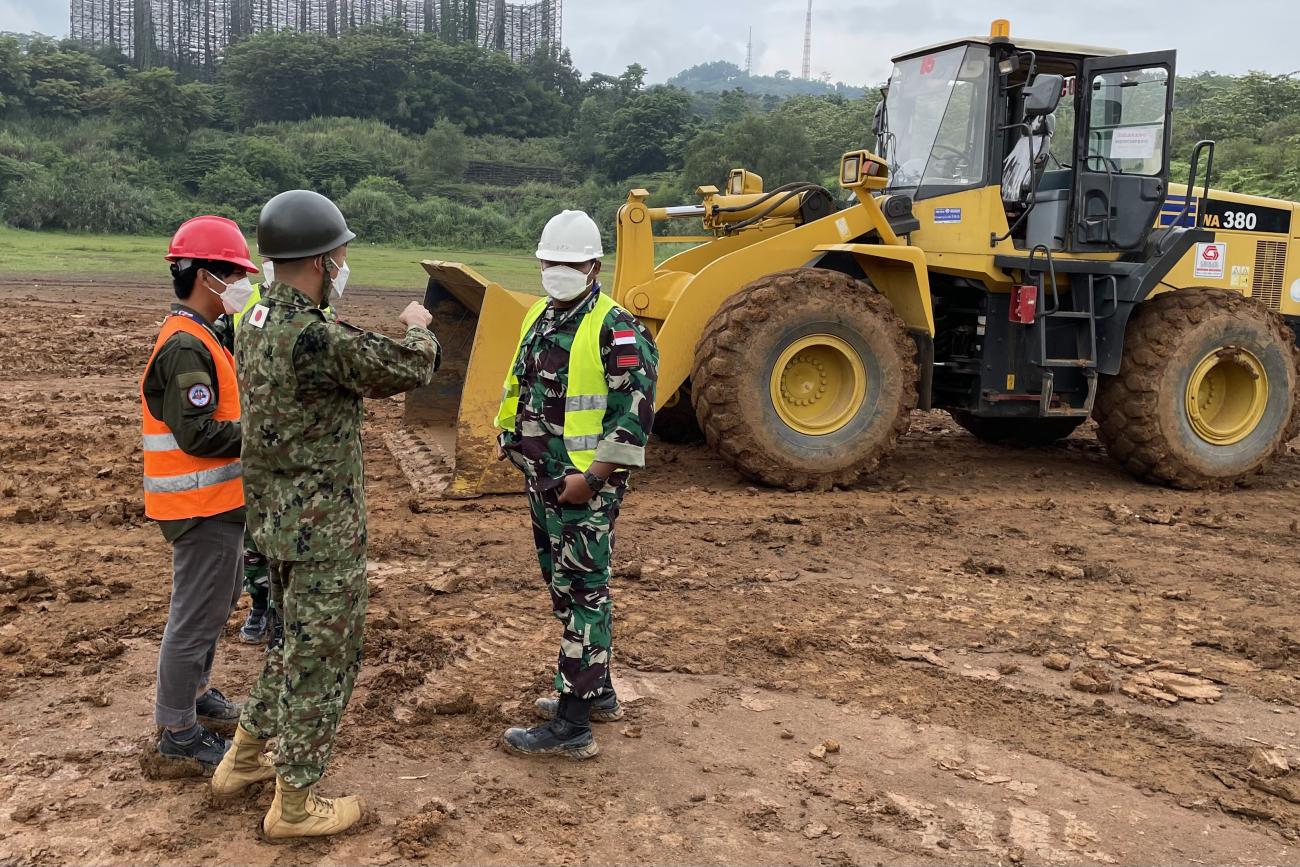 A man wearing an official Indonesian army uniform listens to two men in front of him. Behind them is a yellow bulldozer trying to flat out the land.