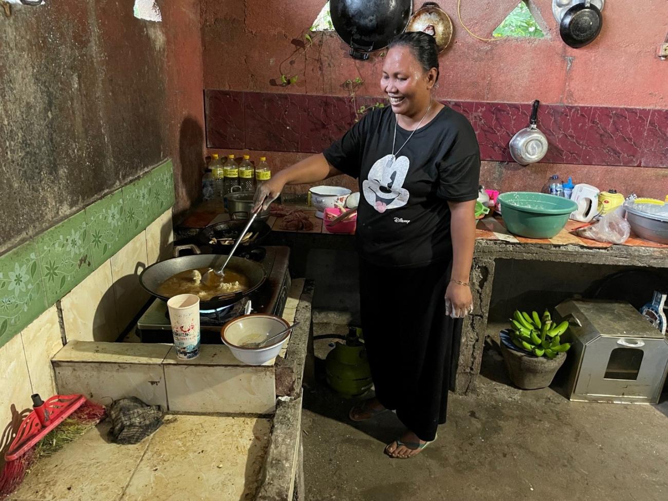 Seorang wanita paruh baya berdiri di dapur ala Indonesia sambil tersenyum saat menggoreng makanan.