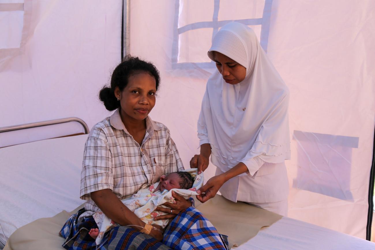 A mother and a newly-born child sits on a bed, a midwife helps with the blanket of the baby
