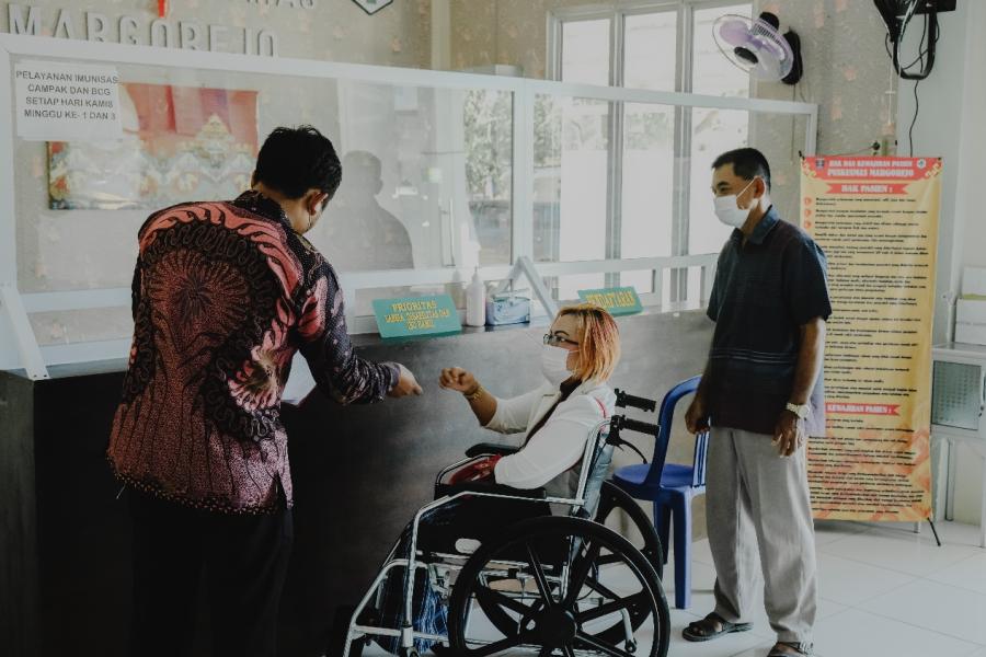Two men assisting a disabled person in a wheelchair to register at the Puskesmas receptionist.