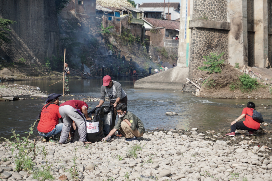 Lima orang membersihkan sungai dan mengumpulkan sampah. Di latar belakang, beberapa tentara Indonesia juga membantu melakukan hal yang sama.