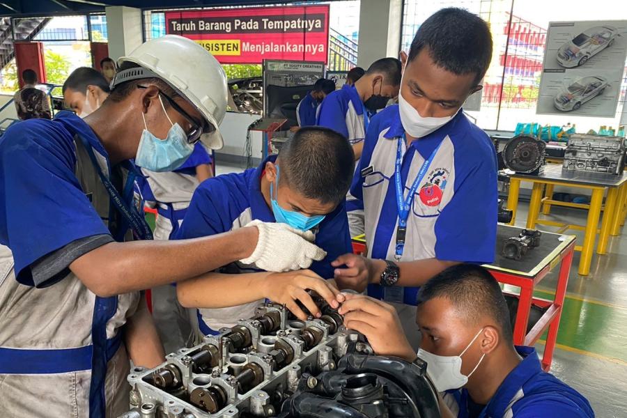 Four vocational students in blue uniforms learn to assemble car engines together as a team.