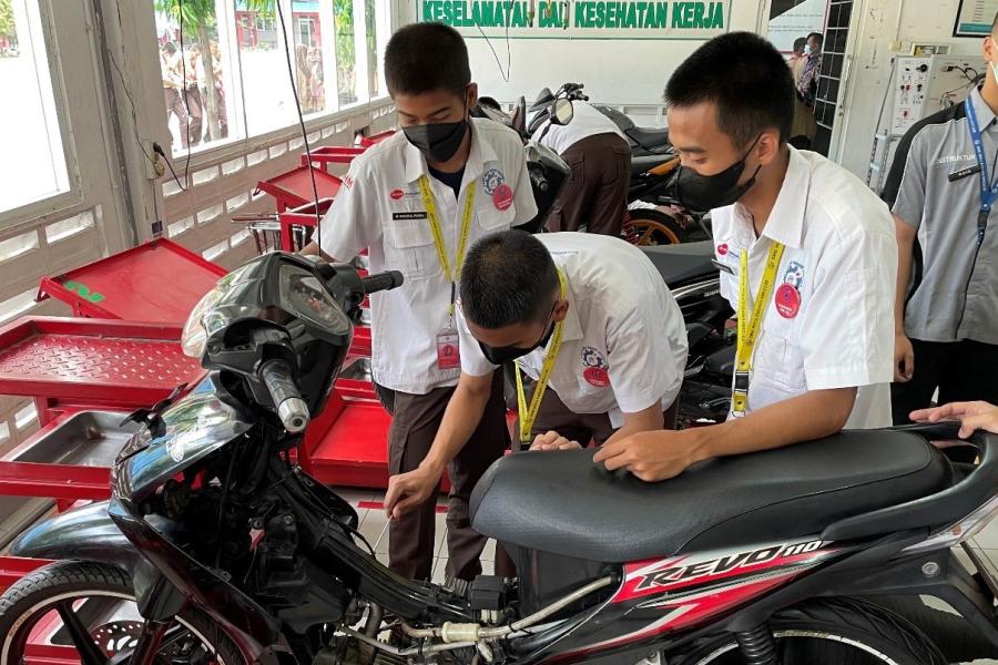 Three male students stand before a black motorcycle as they learn how to fix its engine.