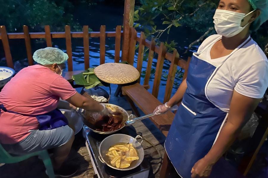 Two middle-aged women prepare local delicacies in a wooden gazebo by the water.