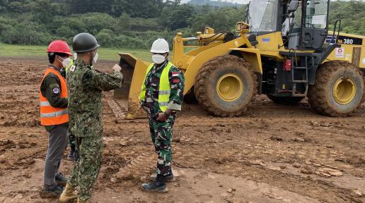 A man wearing an official Indonesian army uniform listens to two men in front of him. Behind them is a yellow bulldozer trying to flat out the land.
